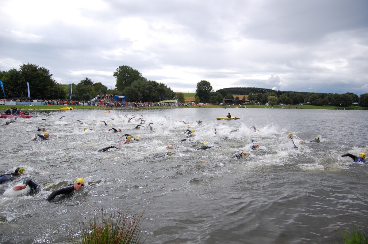 Swimmers in wetsuits and yellow caps racing away from a mass start on a lake, spectators lining the shore