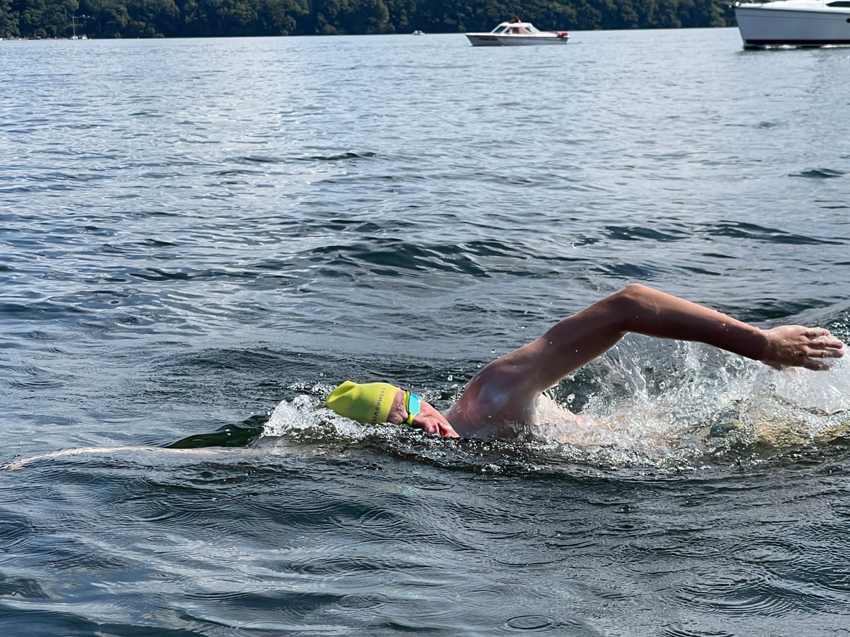 An open water swimmer in a yellow cap swimming front crawl on Windermere, a boat behind