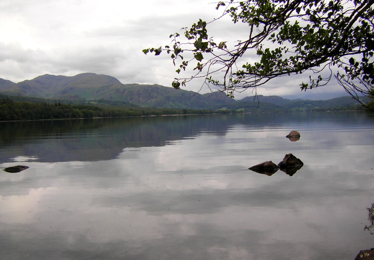 Calm Coniston Water from the shore with the Coniston fells reflected under a grey sky, Lake District