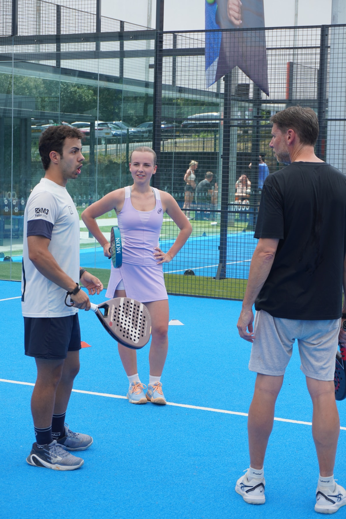 A coach in an M3 Padel Academy shirt talks two players through a drill on a blue padel court